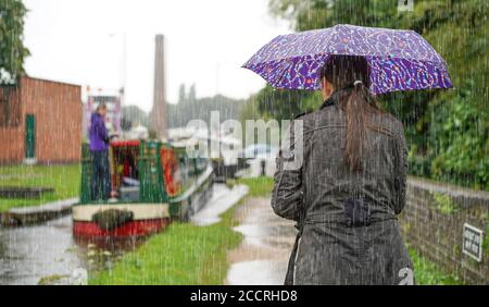 Donna da dietro (corpo superiore) che ripara sotto l'ombrello in piedi sotto la pioggia torrenziale, guardando narrowboat sul canale britannico che si avvicina al blocco canale. Foto Stock