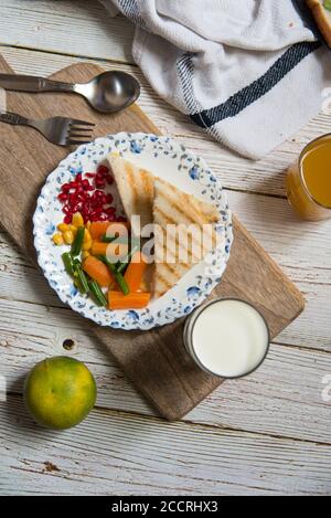 Vista diversa del panino grigliato su un piatto bianco con frutta, latte, condimenti di pane e verdure bollite sullo sfondo Foto Stock