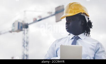 Ritratto di un uomo africano con hardhat utilizzando la tavoletta sul cantiere e osservando il lavoro. Foto di alta qualità Foto Stock