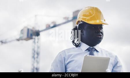 Ritratto di un uomo africano con hardhat utilizzando la tavoletta sul cantiere e osservando il lavoro. Foto di alta qualità Foto Stock