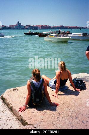 Due turisti a Venezia che si rilassano al sole e si affacciano sull'acqua fino alla Basilica di Santa da un punto dietro Piazza San Marco. Foto Stock