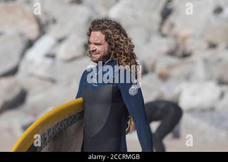 Un surfista maschile con lunghi capelli ricci che porta la sua tavola da surf a Fistral a Newquay in Cornovaglia.