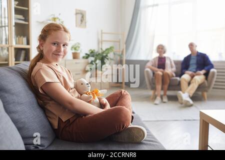 Ritratto dai toni caldi della carina ragazza dai capelli rossi che guarda la macchina fotografica mentre si siede sul divano in un accogliente soggiorno con nonni sullo sfondo, spazio di copia Foto Stock