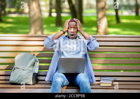Grande fallimento. Giovane ragazzo nero che tiene la testa nel panico vicino al computer portatile al parco Foto Stock