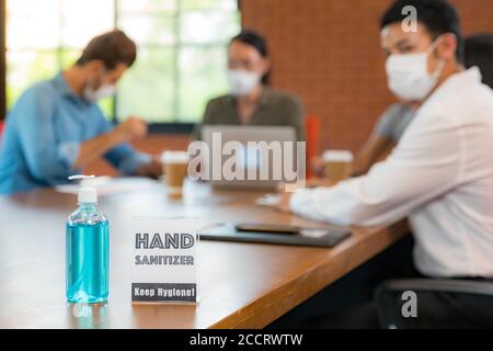 Segnaletica di gel alcolico igienizzante per le mani nella sala riunioni con sfondo di team asiatici che lavorano e si scambiano idee. Indossano una maschera facciale nuova Foto Stock