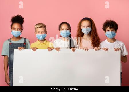 Diversi bambini in maschere facciali con poster vuoto su sfondo rosa, mockup per il design Foto Stock