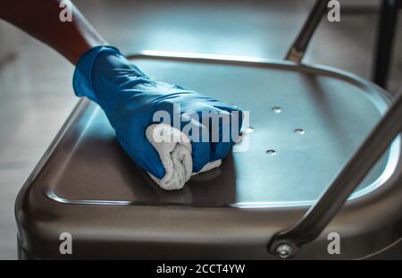 Primo piano della mano di una donna nera che indossa guanti blu e pulisce una sedia. Prevenzione di Covid-19. Messa a fuoco selettiva sulla mano. Sfondo sfocato Foto Stock