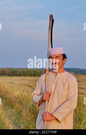 Ritratto di un uomo musulmano sorridente in un cappuccio ricamato e abiti tradizionali bianchi con scythe a mano in un hayfield. Foto Stock