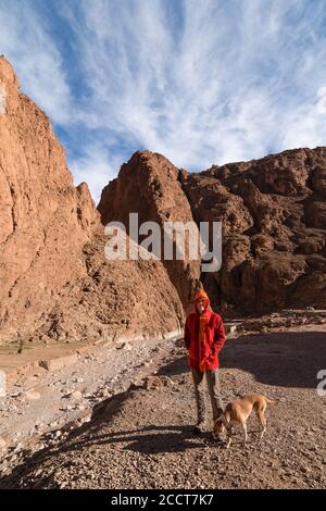 Un uomo incappucciato con una djellaba rossa e il suo cane nella gola di Dades in Marocco. Concetto di viaggio Foto Stock