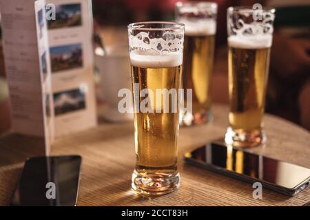 Un tavolo di legno in un pub o bar con 3 bicchieri di birra schiumosa color oro e 2 telefoni cellulari. Nessuna persona visibile Foto Stock