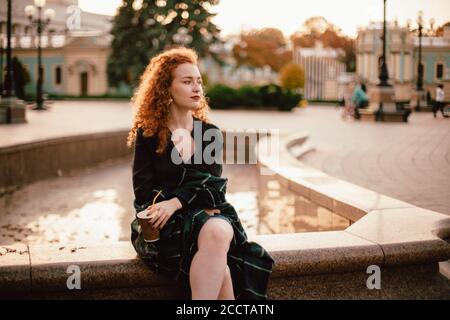 Giovane donna testa rossa pensierosa seduta sulla fontana in città Foto Stock