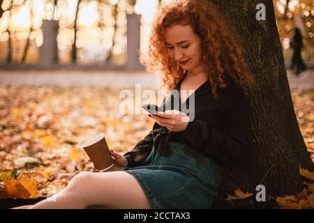 Felice giovane donna che usa lo smartphone mentre si siede da albero nel parco in autunno Foto Stock