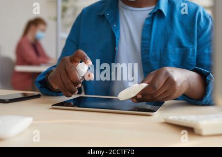 Primo piano di un giovane uomo afroamericano che pulisce compresse con salviette igienizzanti mentre si lavora alla scrivania in ufficio post pandemico, copia spazio Foto Stock