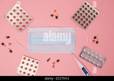 Vista dall'alto verso il basso su una posa piatta di maschera facciale, pillole e termometro su sfondo rosa, concetto di covid e influenza, spazio di copia Foto Stock