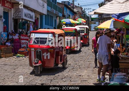 Una linea di tuk tuk o mototaxis passa dal mercato aperto a San Pedro la Laguna, Guatemala. Molte delle donne maya della città indossano ancora i traditi Foto Stock