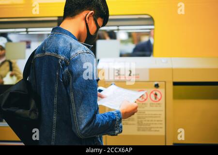 Il giovane indossa una maschera protettiva per il viso mentre apri la mappa e alla ricerca dei luoghi per viaggiare in metropolitana dal retro visualizza Foto Stock