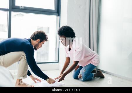 Interracial giovane coppia di sposi si siedono sul pavimento del loro affittato appartamenti, occupato con la composizione di relazione finanziaria per il servizio di assistenza fiscale, utilizzando p Foto Stock