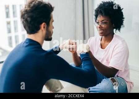 Il ragazzo del Caucaso e la sua ragazza africana pratica yoga, sedersi gambe incrociate, tenendo le mani insieme, guardando ogni altro durante la seduta in legno Foto Stock