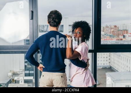 Positiva donna africana abbraccia con il suo bel ragazzo europeo bruna, rilassarsi a casa, dare un caldo abbraccio l'uno all'altro, stare vicino finestra backgr Foto Stock