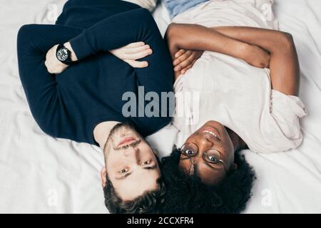 Ritratto di amici felici e diversi che giacciono in un abbigliamento casual con le mani ripiegate l'una accanto all'altra sul letto e guardando la macchina fotografica. Capovolto, vista dall'alto. Foto Stock