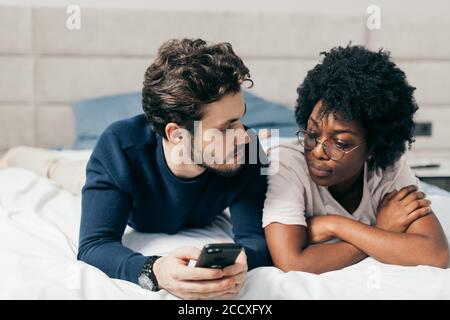 Due amici multirazziali diverti, uomo e donna godono di riposare insieme a letto, hanno espressioni positive, fare selfie foto, condividere immagini in sociale n Foto Stock