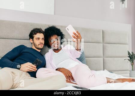 Allegro giovane razza mista coppia amorevole sdraiato a letto e. fare selfie su sfondo grigio chiaro Foto Stock