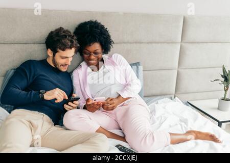 Giovane moglie e marito si rilassano a letto durante la giornata. Spauses di diverse nazionalità che condividono storie divertenti e guardano video con Smart Foto Stock