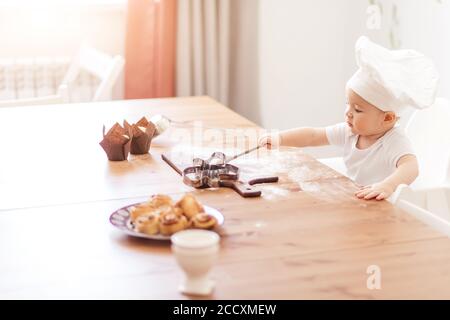 Adorabile bimbo piccolo, 1-2 anni, che indossa il cappello Cook che gioca con le teglie dei muffin, seduto al tavolo coperto di farina e i biscotti serviti. Foto Stock