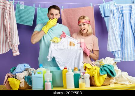 Donna emozionale e uomo in T-shirt essere sorpresi al punto, primo piano ritratto. Isolato sfondo blu. Famiglia trovare macchia sulla T-shirt bianca. Lavanderia Foto Stock