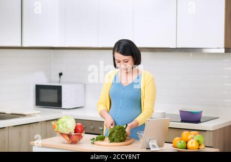 Donna incinta che taglia broccoli per insalata verde fresca, femmina prepara gustosa cena biologica a casa Foto Stock