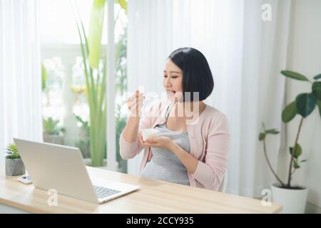 donna incinta sorridente che mangia yogurt mentre si siede a tavola con un computer portatile a casa Foto Stock