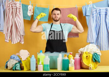 uomo serio bello in grembiule nero, t-shirt blu, guanti gialli che mostrano bicipiti mentre si trova dietro il tavolo isolato su sfondo giallo. hobby, Foto Stock