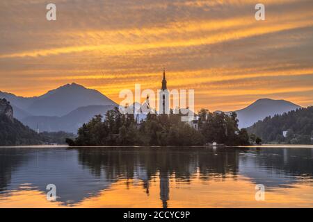 Il lago di Bled con la chiesa di Santa Maria e le montagne sullo sfondo arancione sotto il cielo mattutino, Slovenia, Europa Foto Stock