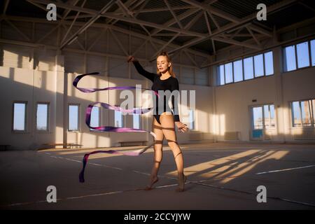 Giovane bella donna in leotard nera che ondola lungo nastro colorato in aria, elemento di ginnastica artistica, concetto sportivo professionale, Foto Stock