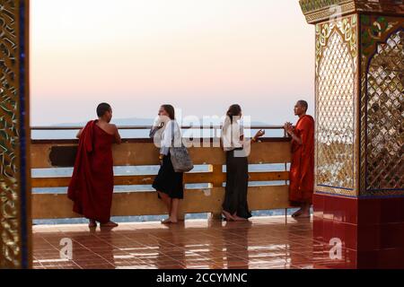 Giovani monaci buddisti vestiti di abiti rossi tradizionali che parlano con le donne turisti al tramonto. A piedi nudi. Su Taung Pyae Pagoda, Mandalay, Myanmar Foto Stock