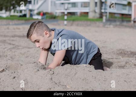 Primo piano di un bel ragazzo che gioca nella sabbia sulla spiaggia, ha un'espressione sorpresa e candida sul volto Foto Stock