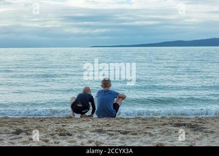 Due giovani fratelli seduti sulla spiaggia che guardano il mare/oceano in pace Foto Stock