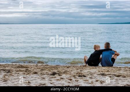Due fratelli si legano sulla bellezza dell'oceano mentre si siedono sulla spiaggia, con il fratello minore che conforta il più grande con un braccio intorno a lui. Foto Stock