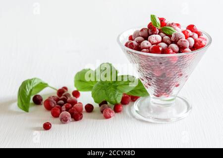 Mirtilli congelati in un recipiente di vetro su una tavola bianca, bacche e foglie di basilico verde. Cibo surgelato, concetto di conservazione. Piante naturali di curry. Foto Stock