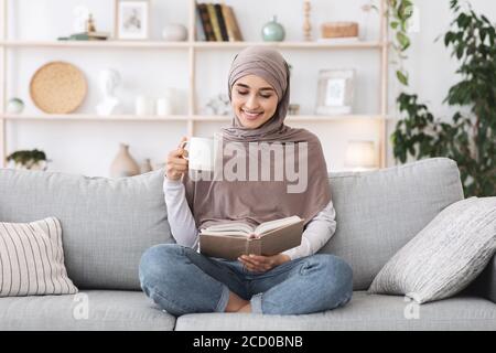 Tempo libero preferito. Libro di lettura rilassato della ragazza musulmana e tè bevente a casa Foto Stock