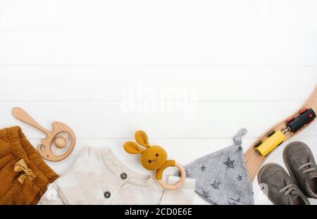 Vestiti per bambini, stivaletti e giocattoli in legno su sfondo di legno bianco con spazio vuoto per il testo. Vista dall'alto, disposizione piatta. Foto Stock