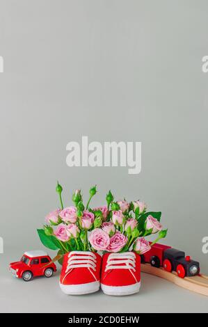 Paio di scarpe rosse per bambini piene di fiori di rosa e giocattoli su sfondo grigio. Spazio vuoto per il testo. Foto Stock