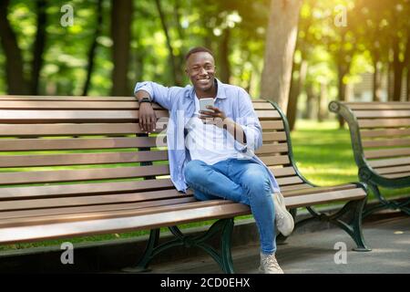 Uomo nero sorridente con telefono cellulare rilassante sulla panca a. parcheggia il giorno d'estate Foto Stock