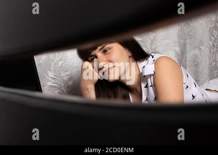 Una bambina di brunette si trova sul letto e guarda il film sullo schermo del computer portatile. Riposo dopo il lavoro Foto Stock