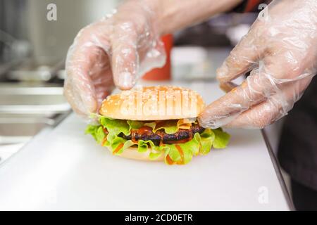 Le mani dello chef dimostrano il Burger finito. Preparazione dell'hamburger al ristorante. Mani del cuoco in guanti monouso in base ai requisiti di igiene Foto Stock