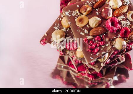 Barrette di cioccolato spezzate fatte a mano decorate con nocciole intere, mandorla e lampone sublimato su sfondo rosa con riflessione; fuoco selettivo, bl Foto Stock