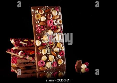 Barretta di cioccolato fatta a mano decorata con nocciole intere, mandorla e lampone sublimato su sfondo nero con spazio vuoto per il testo; fuoco selettivo Foto Stock