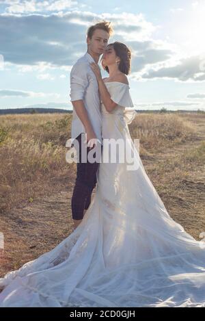 giovane coppia di sposa e sposo in abito da sposa, vestito bianco, sul campo, sullo sfondo del cielo blu nuvoloso. Tiro al matrimonio, sposa An Foto Stock