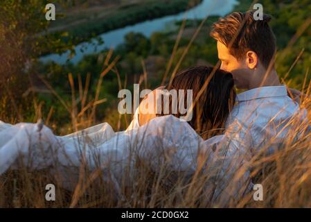 mezzo ritratto di una bella coppia di ragazzo e ragazza sdraiata in erba. Sullo sfondo del sole che tramonta in una serata estiva. Abito bianco di Th Foto Stock