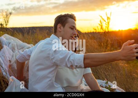 giovane coppia di sposi e sposi in un campo erboso, prendere un selfie da uno smartphone. Abito da sposa bianco. Il ragazzo nel vestito bianco. Coppia amorevole taki Foto Stock